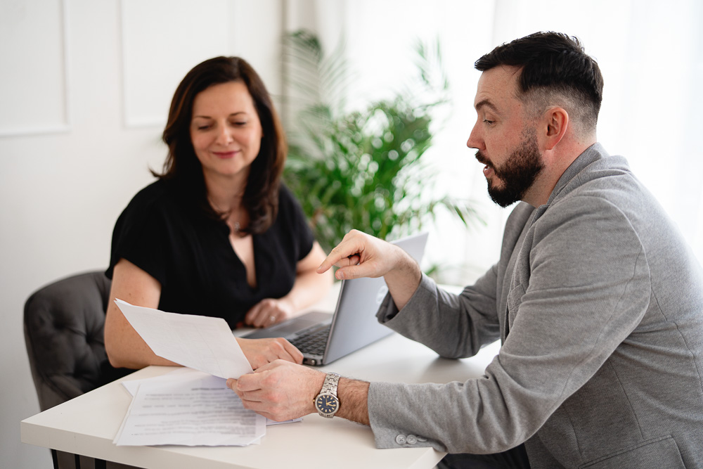 Muž ukazuje na dokument, zatímco žena sedí s notebookem u stolu v kanceláři.