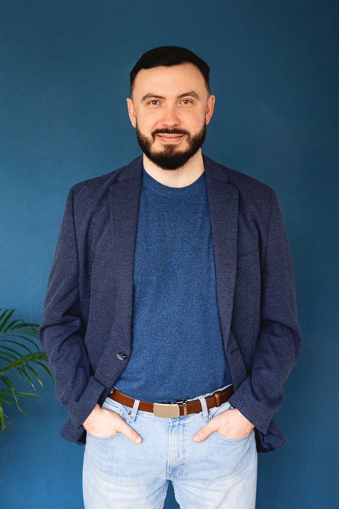 Muž s tmavými vlasmi a plnovousem stojí před modrým pozadím, má modré tričko, tmavě modré sako a světlé džíny s hnědým opaskem.