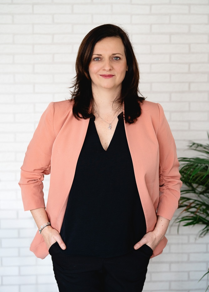 Žena s tmavými vlasy v černém topu a růžovém saku stojí před bílou cihlovou stěnou s rukama v kapsách.