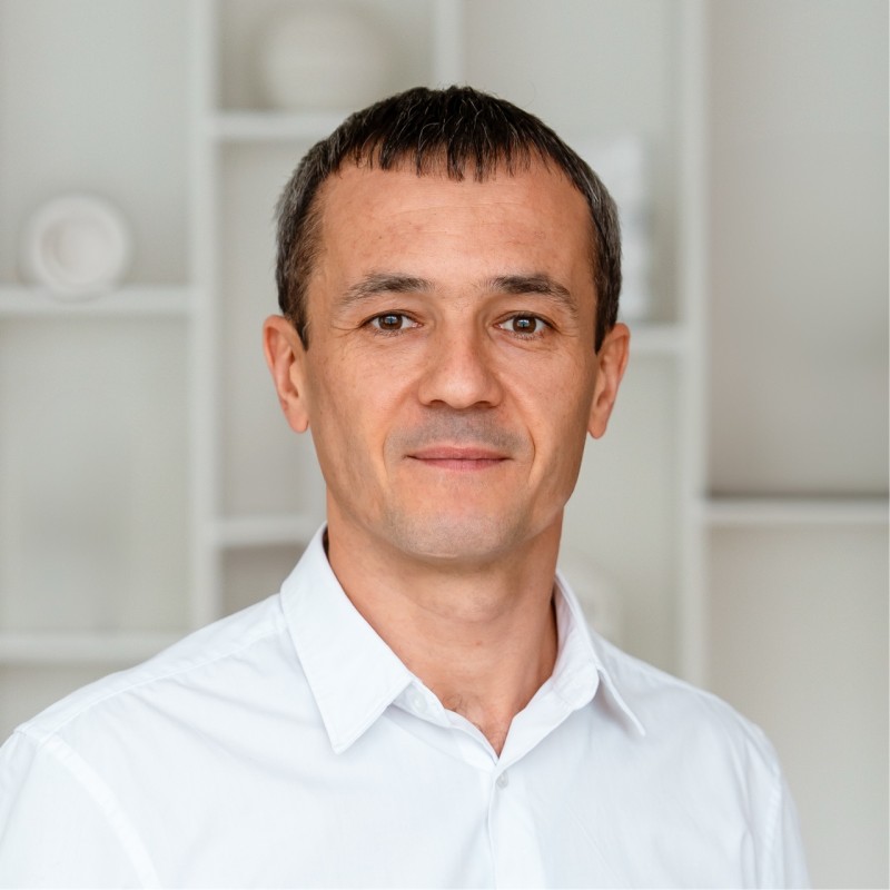 Muž s krátkými vlasy, jemným vousem a pihami na tváři, oblečený v červeném tričku, pózuje před bílou stěnou.