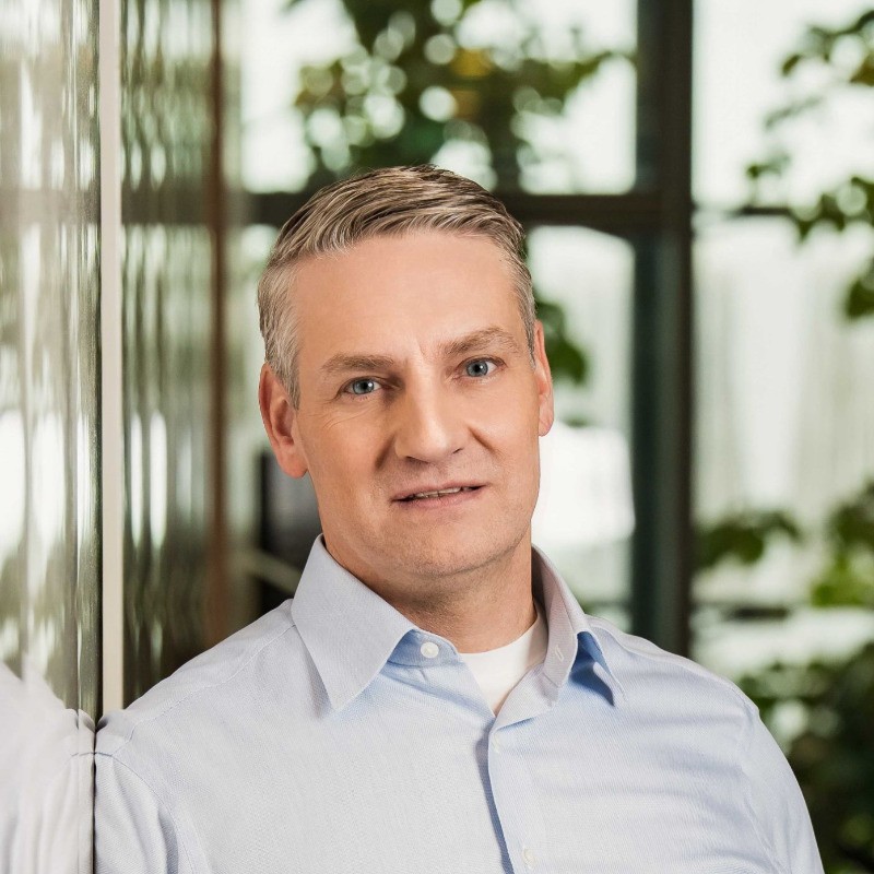 Usměvavá žena s dlouhými světlými vlasy v culíku, oblečená v zelených květovaných šatech, stojící venku s rozostřeným městským pozadím.