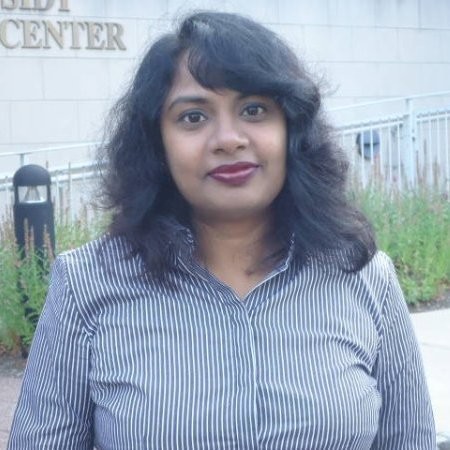 Žena s hnědými vlasy se usmívá venku, v pozadí jsou stromy a žluté listí.