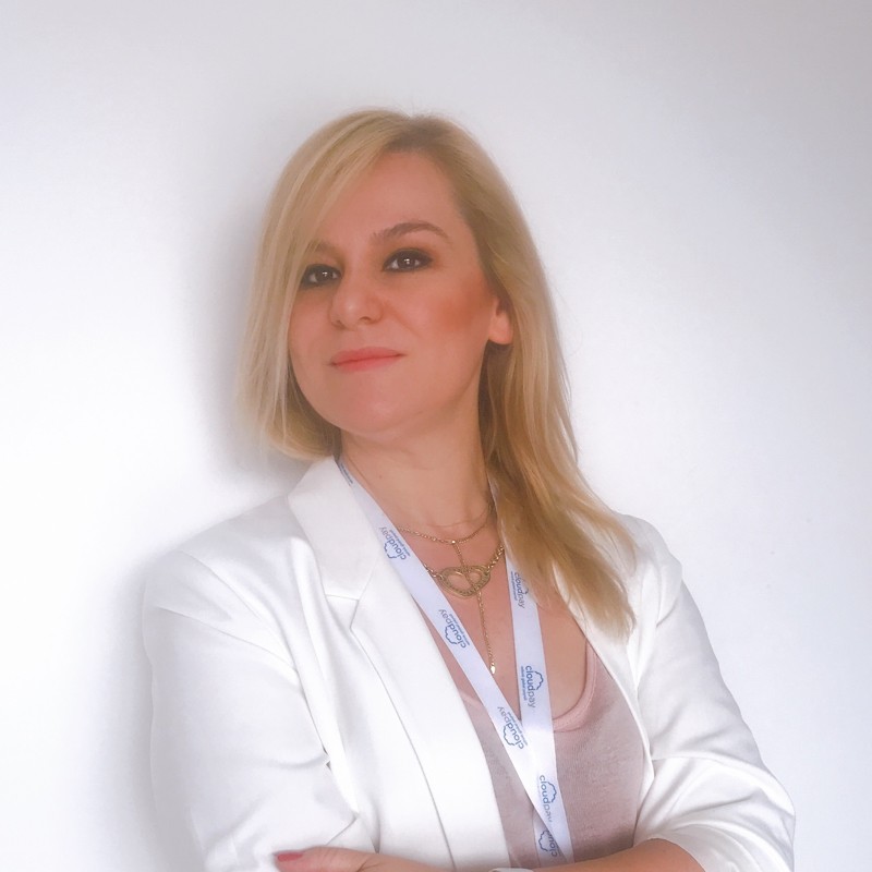 Žena s hnědými vlasy se usmívá venku, v pozadí jsou stromy a žluté listí.