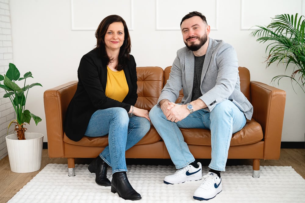 Muž a žena sedí na hnědé sedačce v moderní místnosti s rostlinami.