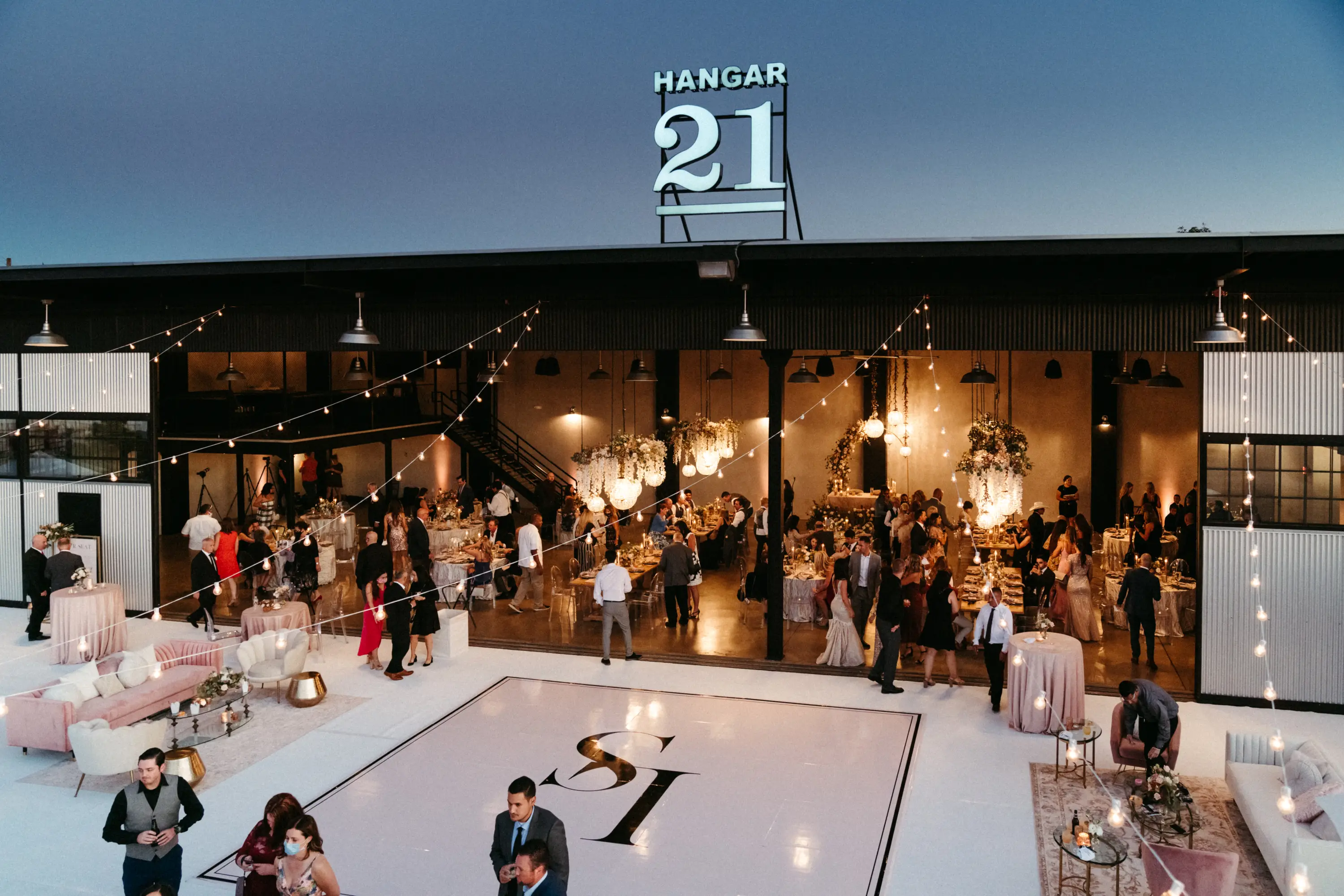 Evening event at Hangar 21 with guests mingling inside and outside a decorated venue featuring string lights, floral arrangements, and seating areas.