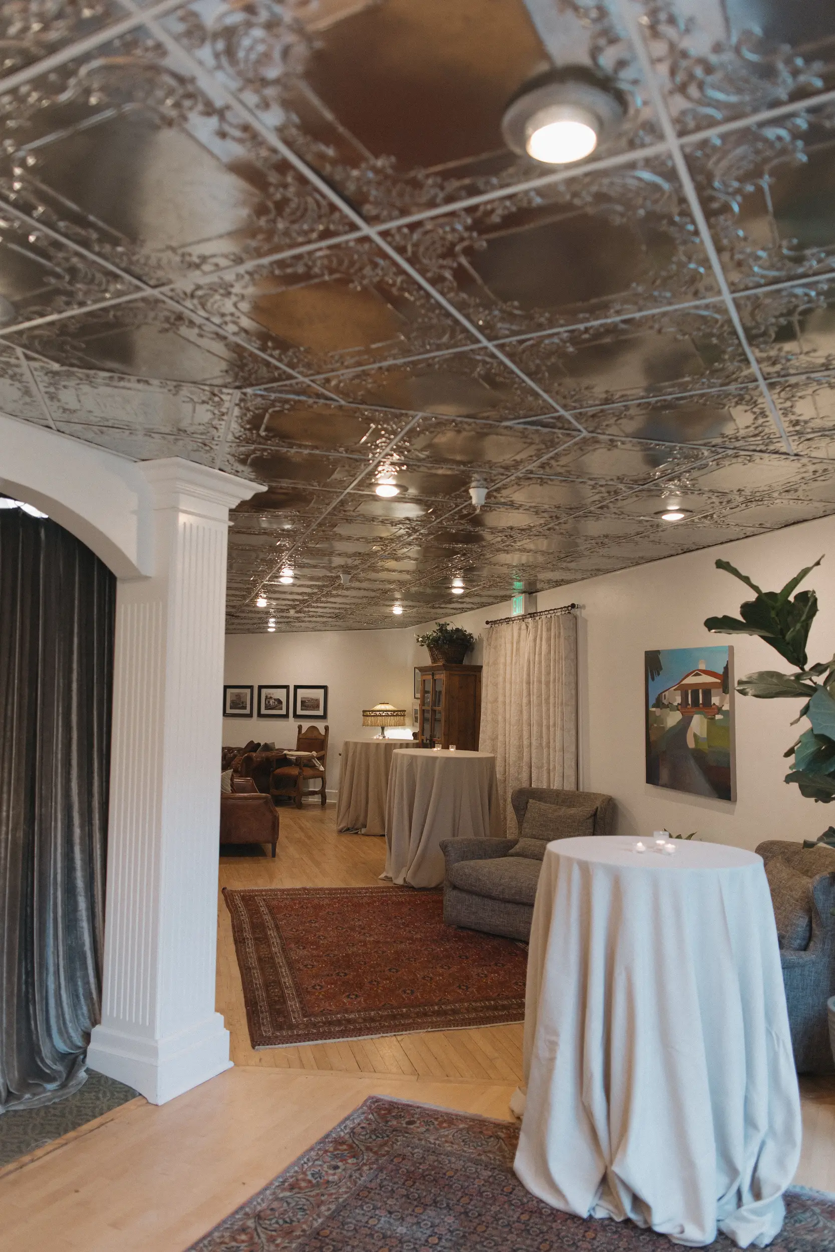 Interior of an elegant event space with ornate ceiling tiles, sofas, tall cocktail tables with white linens, and framed artwork on the walls.