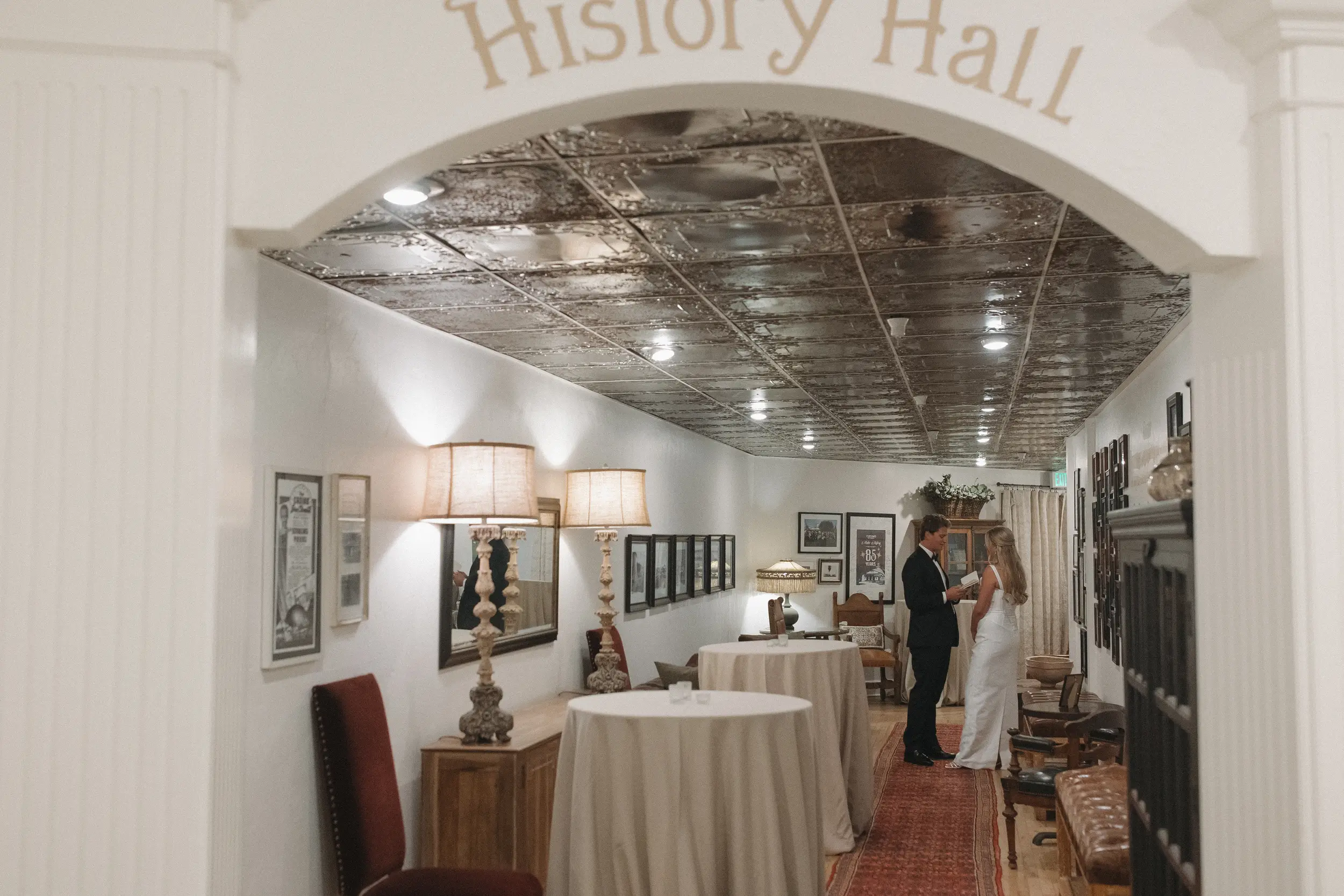 A couple dressed formally standing and talking inside a decorated hallway labeled History Hall, featuring vintage lamps, framed pictures, and covered round tables.