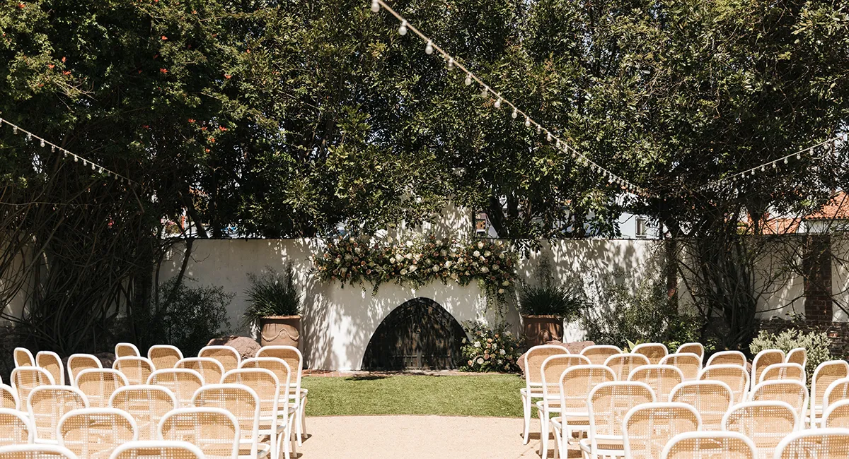 Outdoor wedding ceremony setup with rows of white chairs facing a white wall decorated with an arch of flowers and greenery, surrounded by trees and string lights overhead.