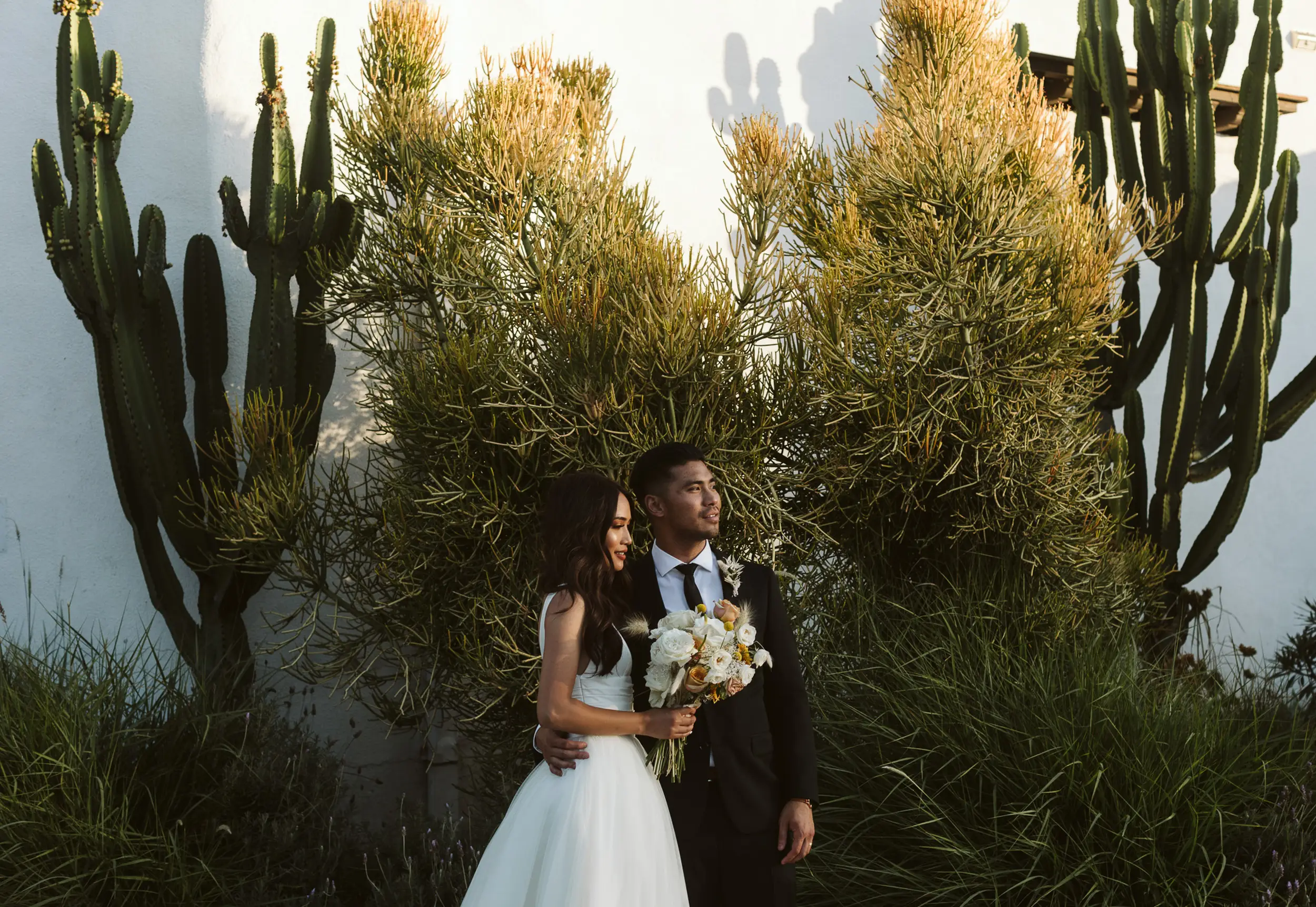 Bride in white gown holding bouquet stands next to groom in black suit in front of tall green cacti and bushes.