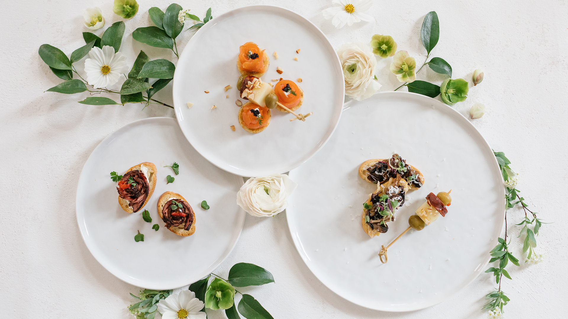 Three white plates with gourmet appetizers, including smoked salmon bites, beef bruschetta, and a cheese and olive skewer, decorated with white flowers and green leaves on a light surface.
