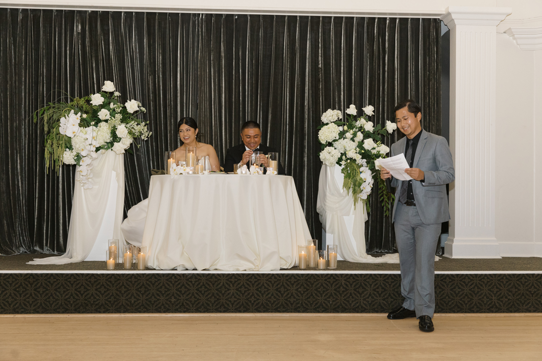 Bride and groom sit at their sweetheart table on stage while a guests reads a toast from the dance floor.