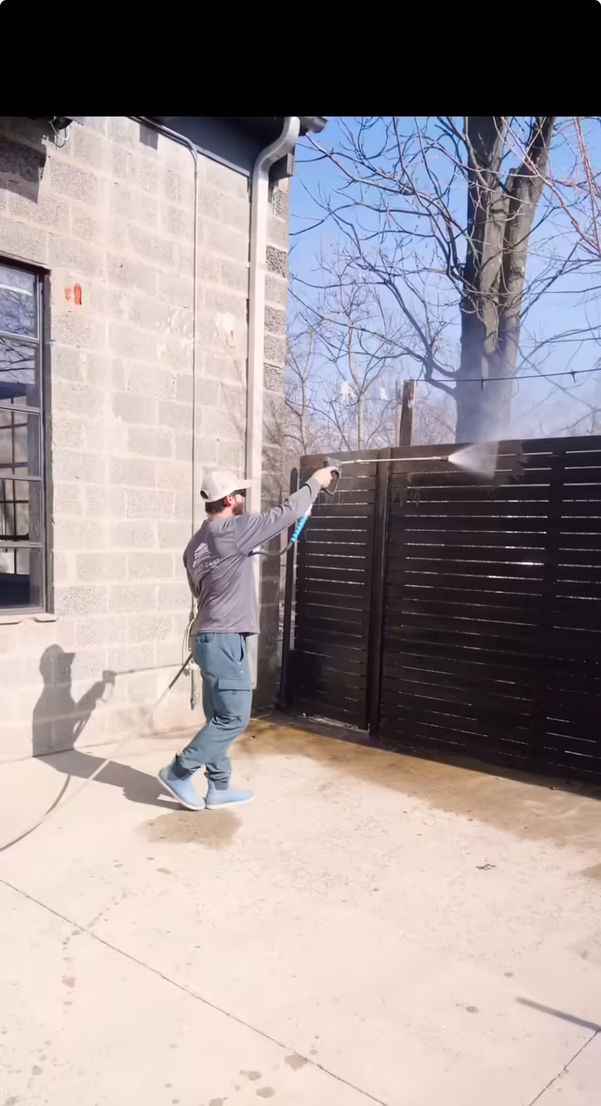 Man wearing a hat and long-sleeve shirt pressure washing a dark wooden fence outside on a sunny day.