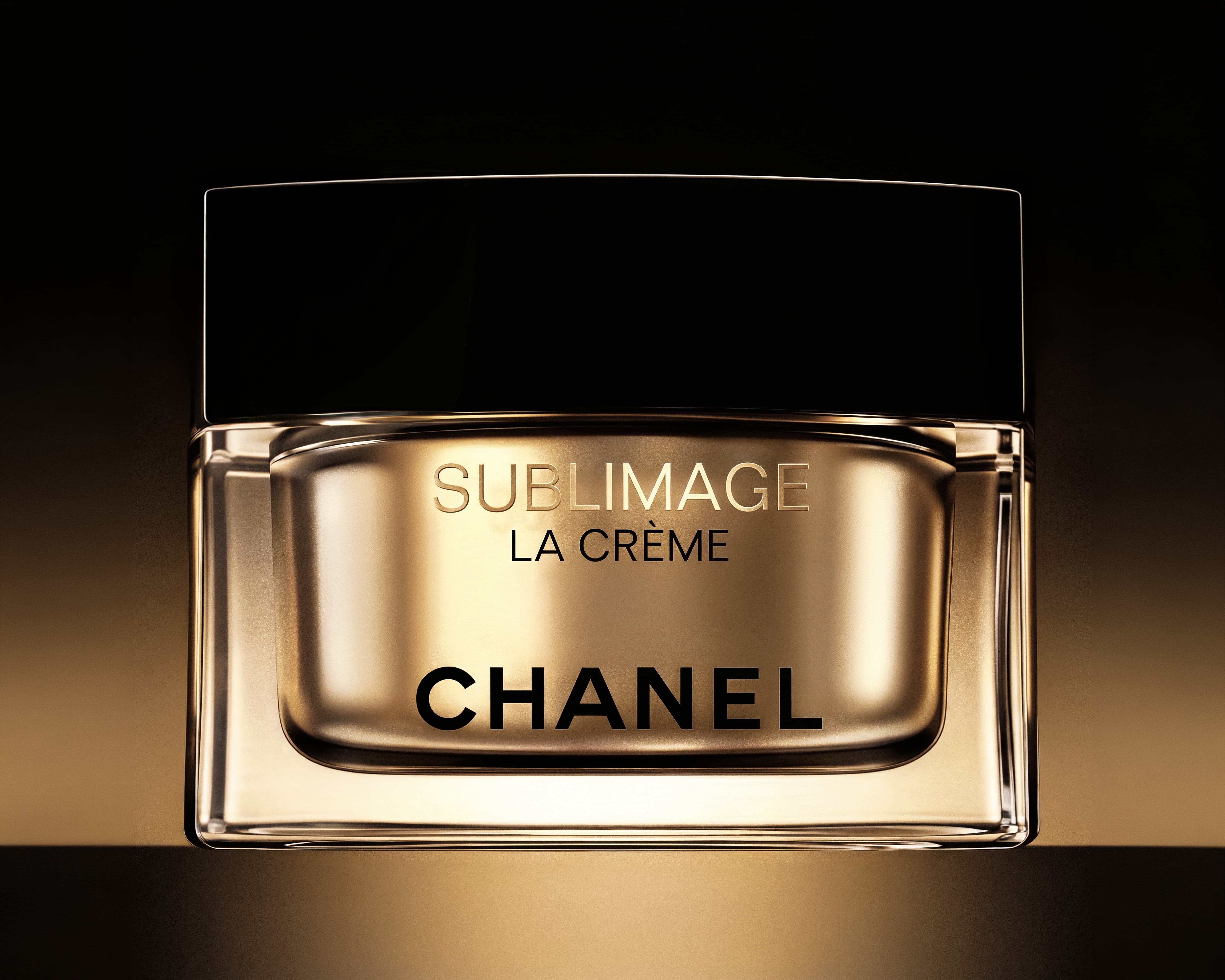 Close-up of a gold and black jar of Chanel Sublimage La Crème skincare cream on a gradient brown background.