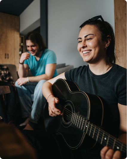Smiling woman playing black acoustic guitar while a man sits and laughs in the background.
