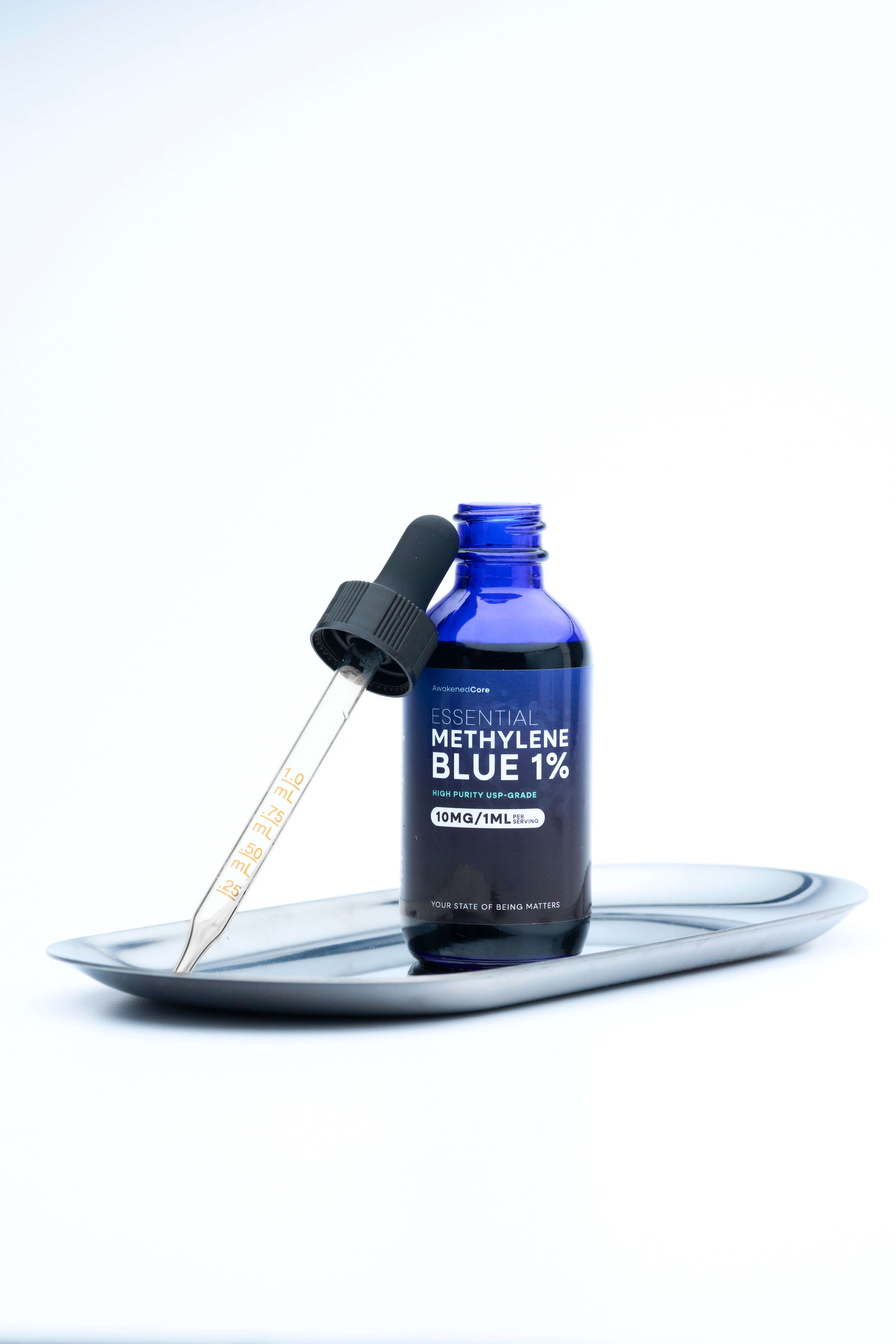 Minimal product photography of an Essential Methylene Blue 1% glass bottle with a graduated dropper resting on a small metal tray, styled against a clean white background with soft studio lighting for a clinical, premium ecommerce presentation.