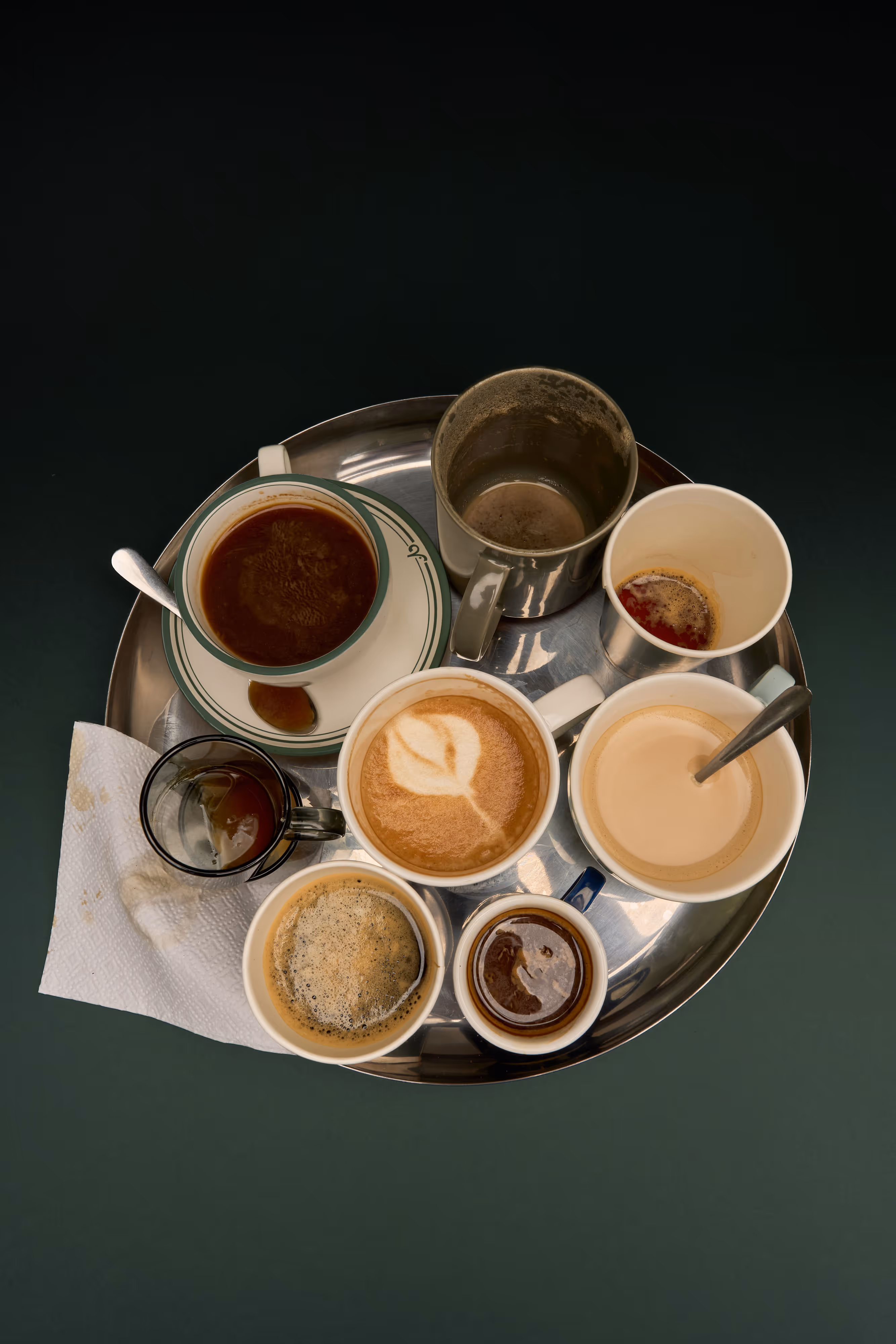 Gooper still life photography for coffee and hospitality brands, featuring an overhead studio composition of assorted coffee cups, espresso, latte art, metal tray styling, and a dark green editorial backdrop.