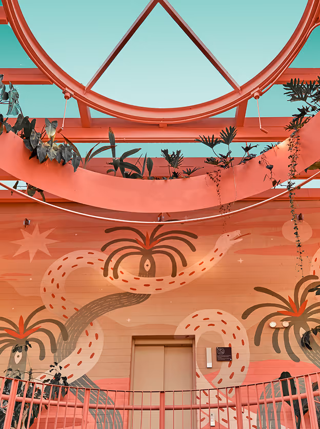Gooper interior and architectural photography for branded spaces, featuring a coral-toned atrium with tropical mural artwork, hanging greenery, glass ceiling structure, and warm daylight styling for hospitality and retail environments.