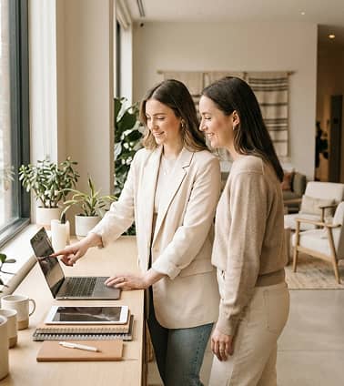 Two women standing by a desk near a window, one pointing at a laptop screen while the other watches, with notebooks and coffee cups on the desk.