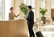 Hotel receptionist assisting a male guest with a suitcase at the front desk in a spacious lobby.