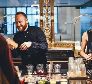Smiling bartender serving drinks in a busy bar with glasses and cocktail ingredients on the counter.