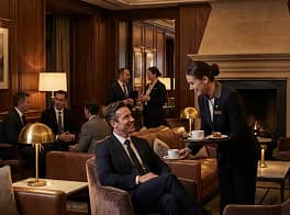 A well-dressed man in a suit sits in a lounge chair smiling as a waitress offers him a cup of coffee in an elegant wood-paneled room.