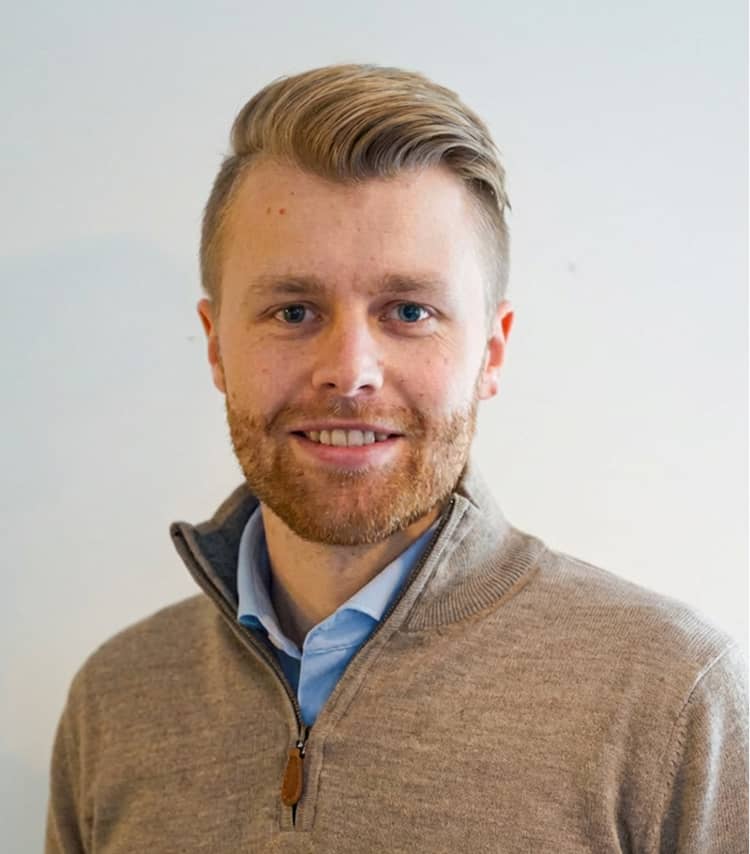 Portrait of a smiling man with short blond hair and beard, wearing a brown zip-up sweater over a blue shirt.