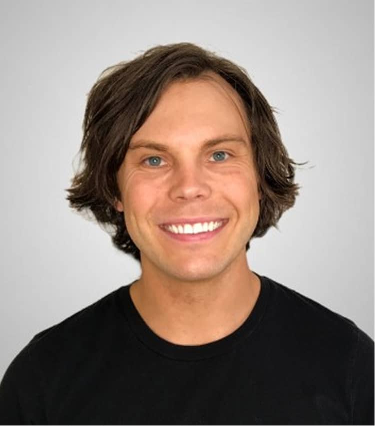 Smiling young man with medium-length brown hair wearing a black shirt against a light gray background.
