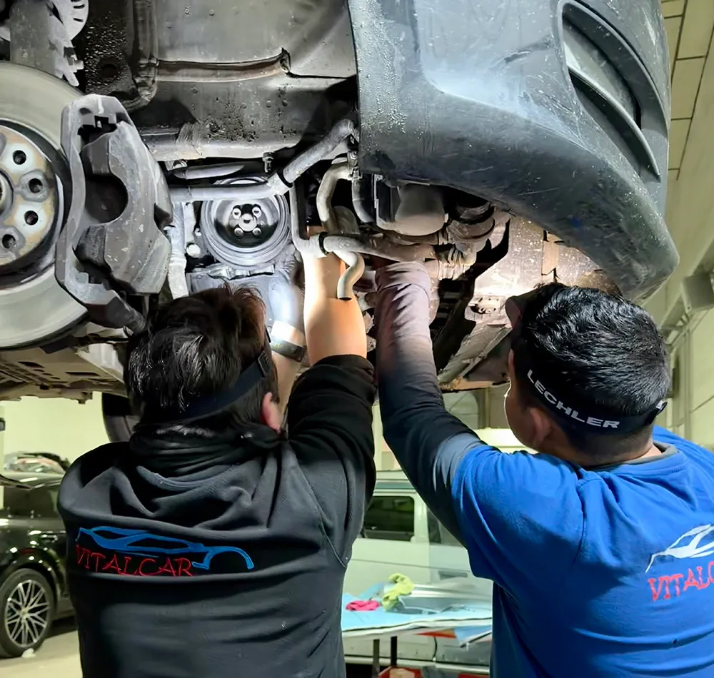 Dos mecánicos trabajan juntos debajo de un coche levantado, inspeccionando el sistema de frenos y suspensión.