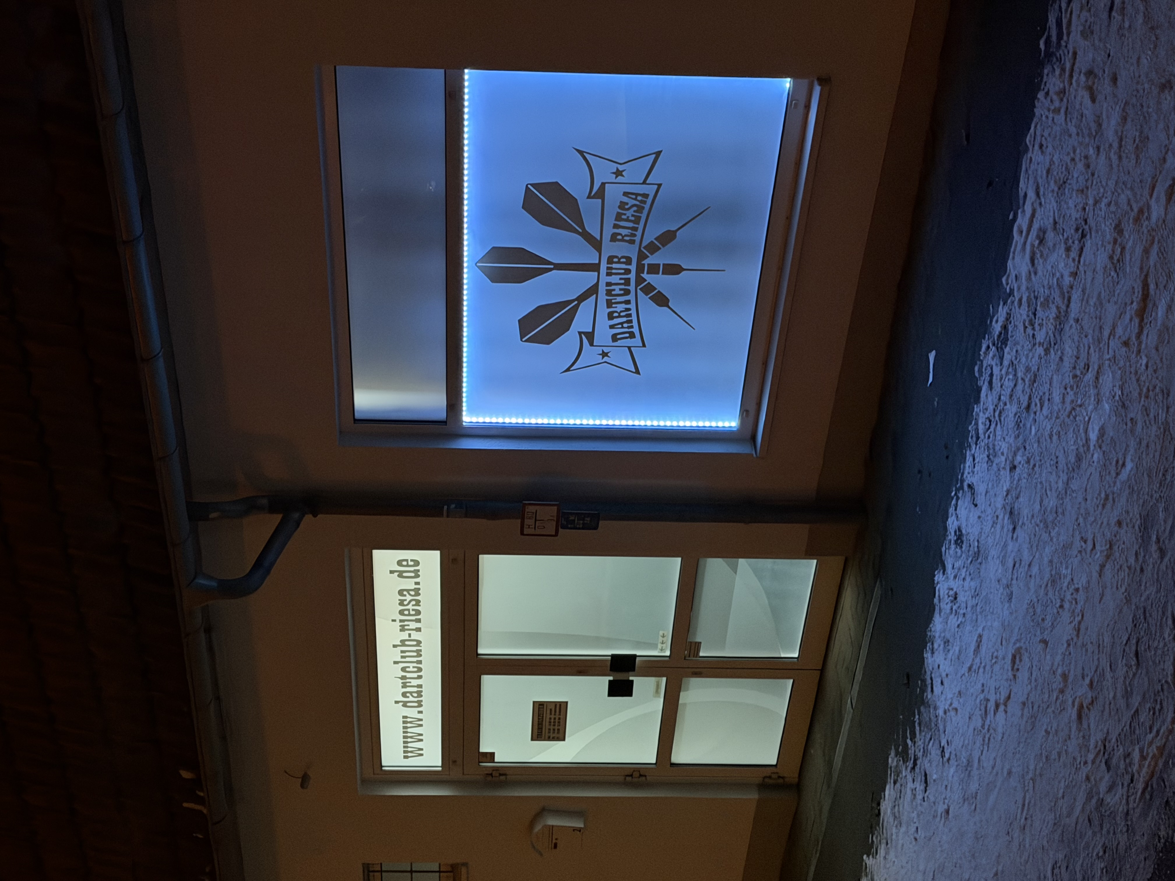 Die Fassade des Dartclub Riesa bei Nacht mit beleuchtetem Vereinslogo neben der Eingangstür.