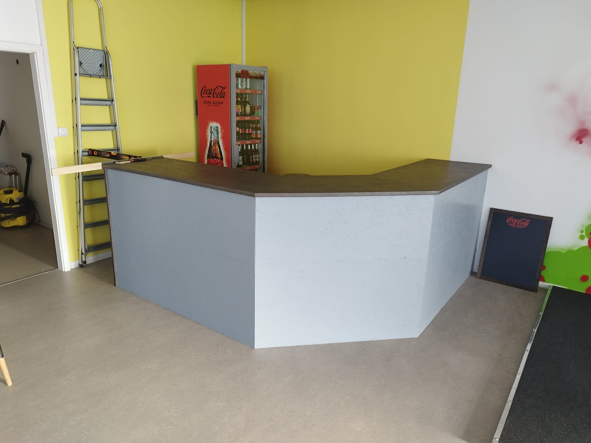 Ein grauer Tresen steht vor einer gelben Wand, daneben befindet sich ein Coca-Cola-Kühlschrank, eine Leiter und ein kleiner Coca-Cola-Tafelaufsatz an der Wand.