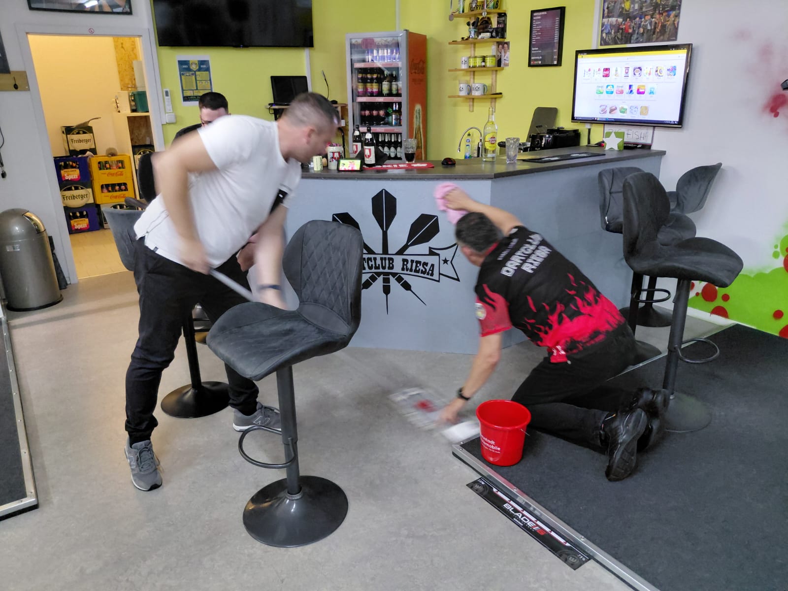 Zwei Männer putzen den Boden vor einer Bar mit dem Logo Dartclub Riesa, einer wischt auf dem Boden und der andere mit einem Besen.