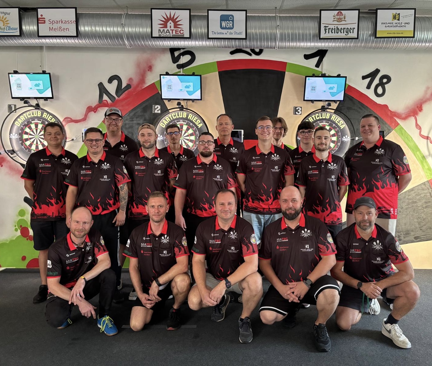Gruppenfoto von 17 Männern in rot-schwarzen Dartclub Riesa Trikots vor Dart-Scheiben an einer Wand mit Sponsorlogos.