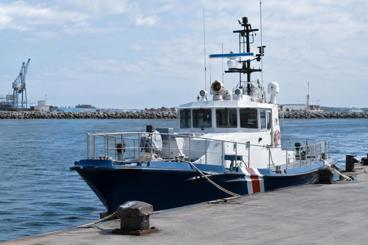 A blue-and-white workboat with a tall mast is moored beside a concrete pier, with harbor water, a rocky breakwater, and cranes in the background.