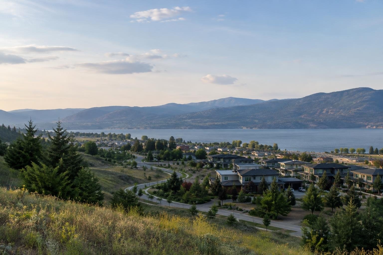 A lakeside neighborhood with houses, roads, and trees lies below a grassy hillside, with a broad lake and forested mountains beneath a partly cloudy sky.