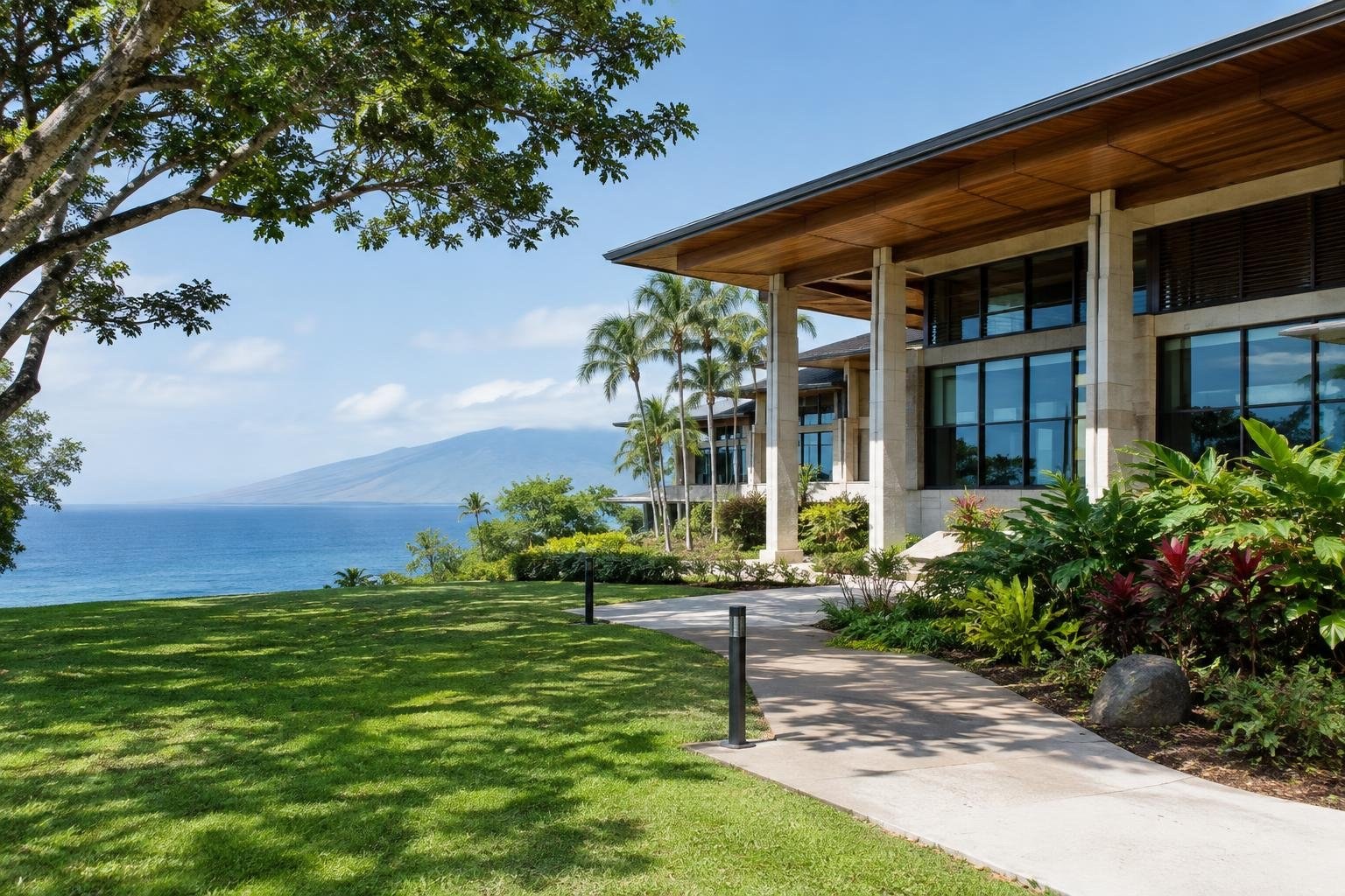 A modern building with broad windows and a wooden roof stands beside a landscaped lawn and path, overlooking the ocean and a hazy mountain.