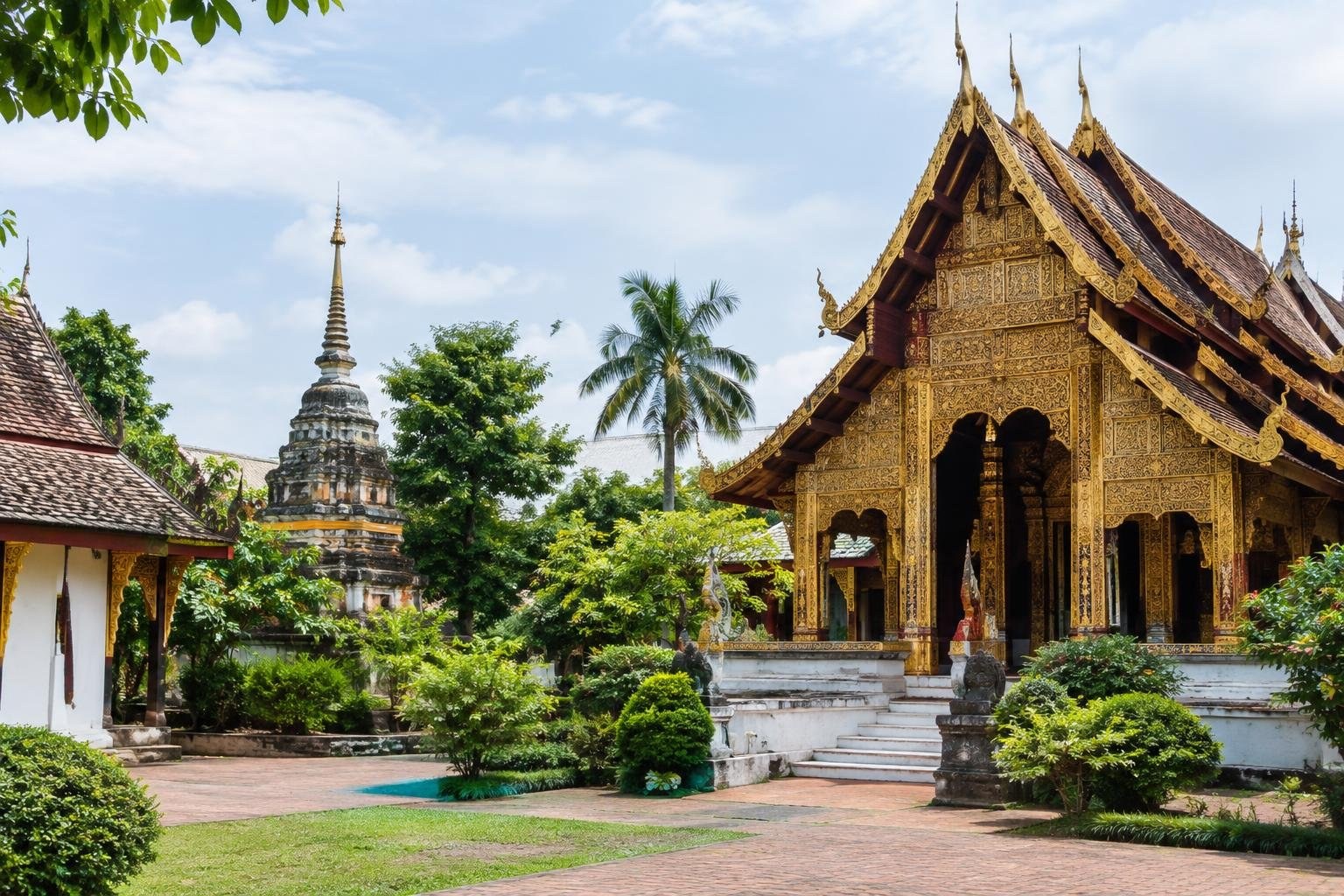 An ornate gold-decorated temple hall and a weathered stone spire stand among tropical trees and shrubs around a paved courtyard.