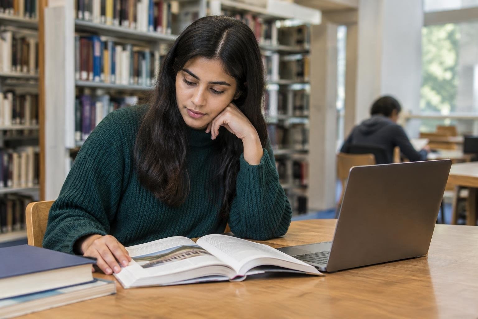 A woman in a dark green sweater reads an open book beside a laptop at a library table, with bookshelves and another seated person in the background.