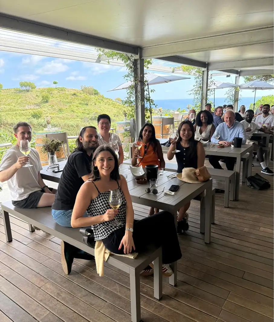 Large group of people enjoying drinks and socializing on a sunny seaside terrace with ocean views.