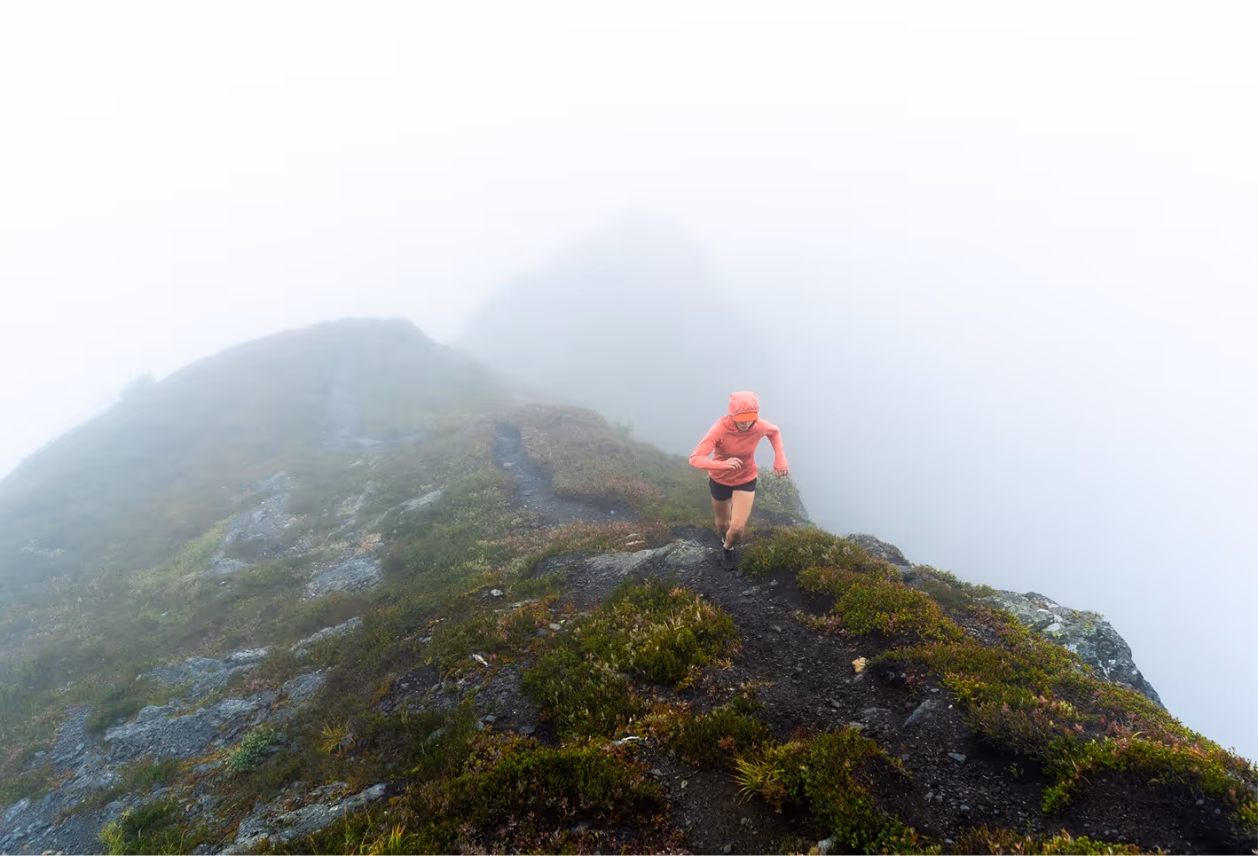 Person in a pink jacket running uphill on a foggy mountain trail surrounded by rocks and greenery.