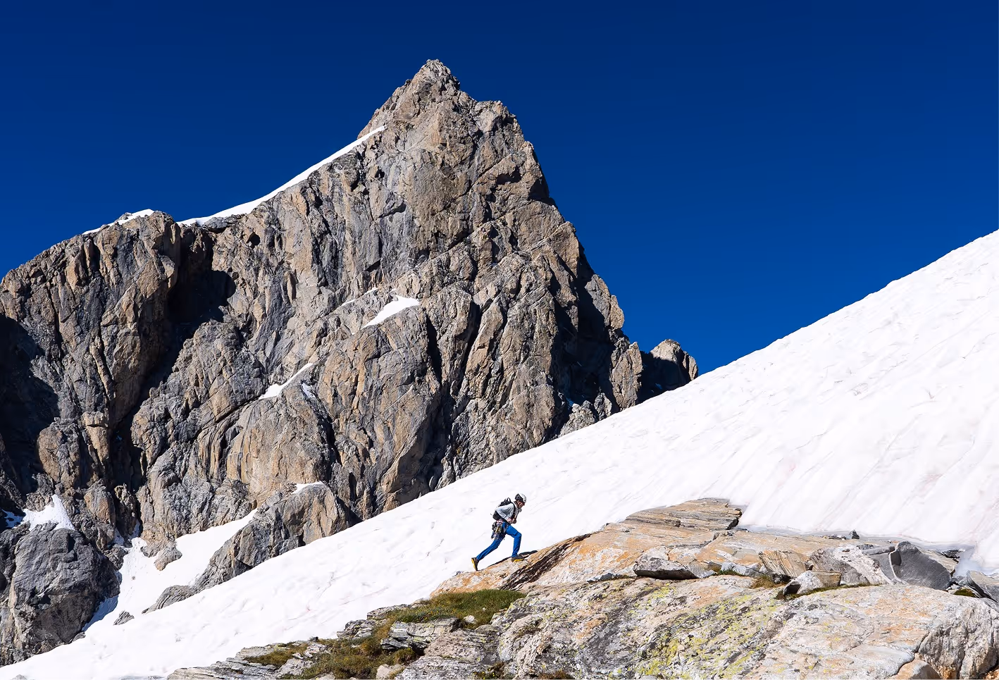 Hiker ascending a rocky slope with snow and a large rugged mountain under a clear blue sky.