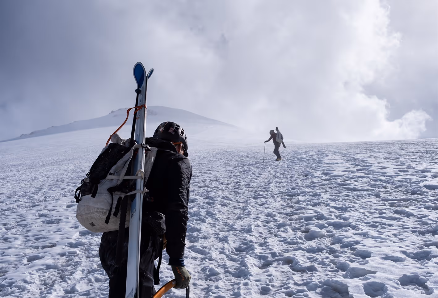 Two skiers climbing a snowy mountain slope under a cloudy sky, one carrying skis and using trekking poles.