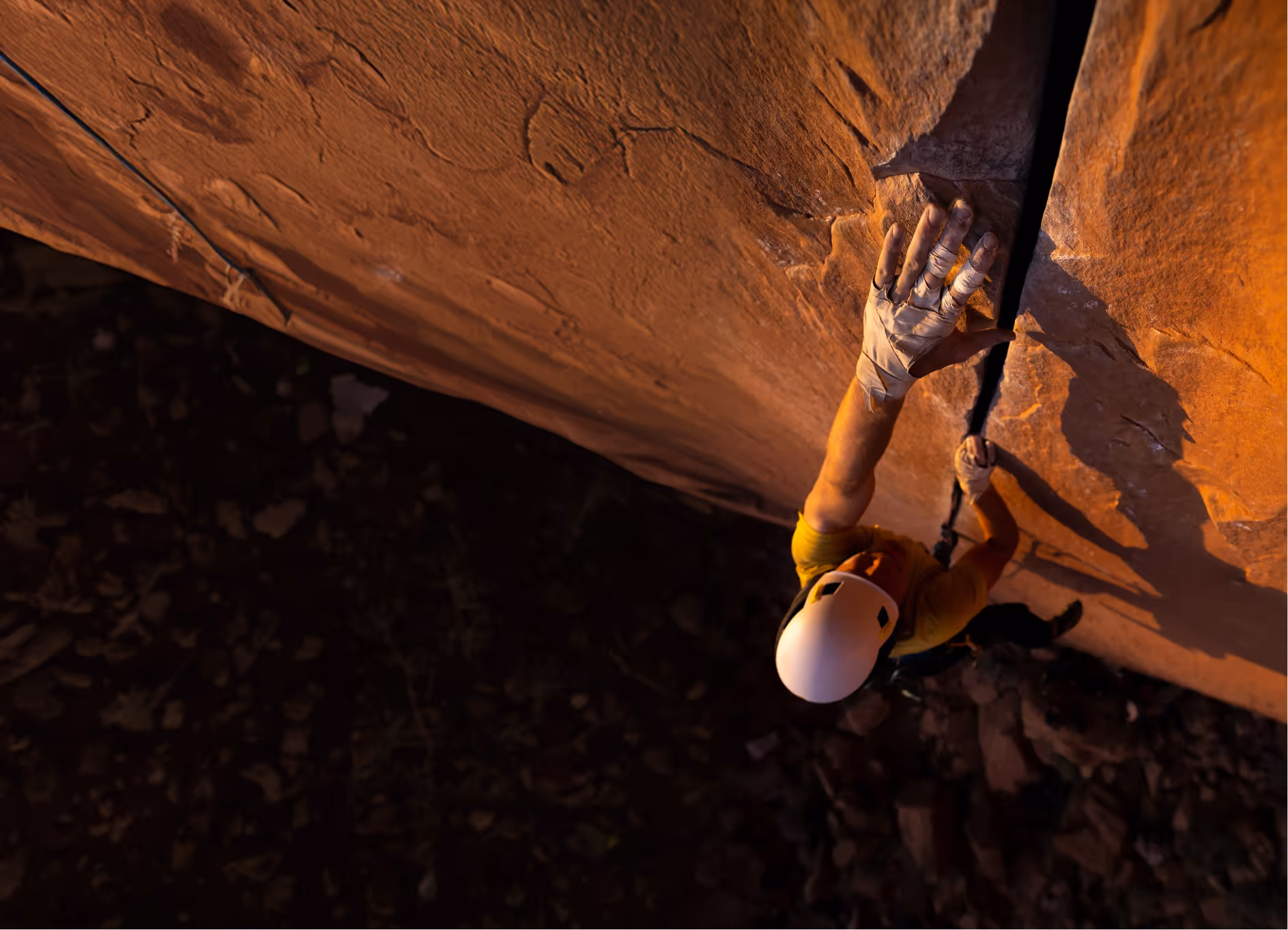 Rock climber wearing a helmet and tape on fingers ascending a vertical rock face with a crack for handhold.