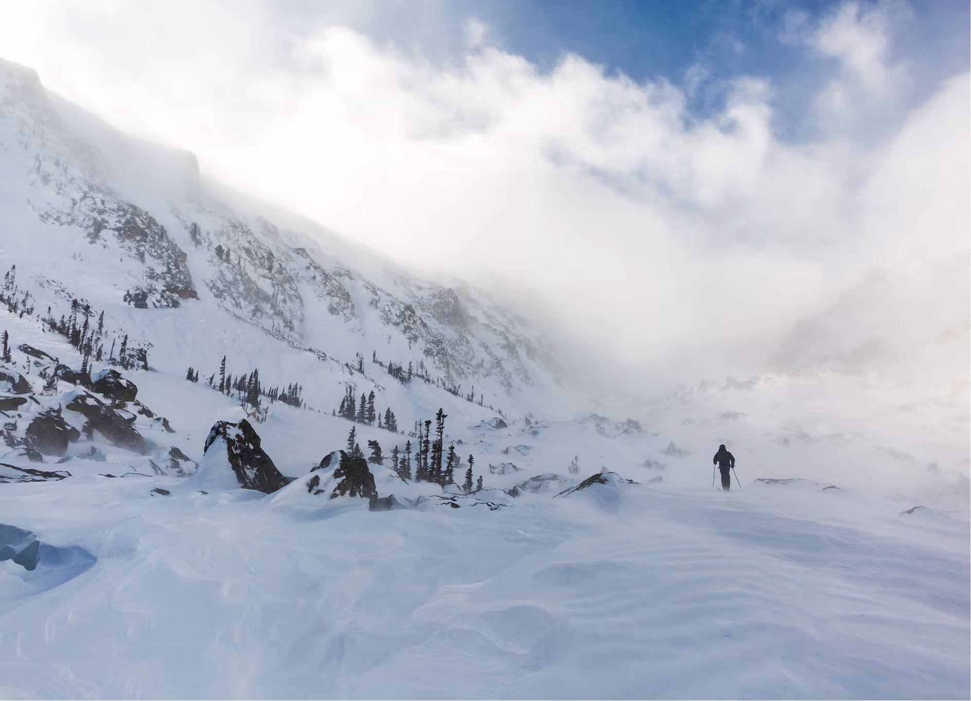 A lone skier ascending a snowy mountain slope surrounded by rocks and sparse trees under a cloudy sky.