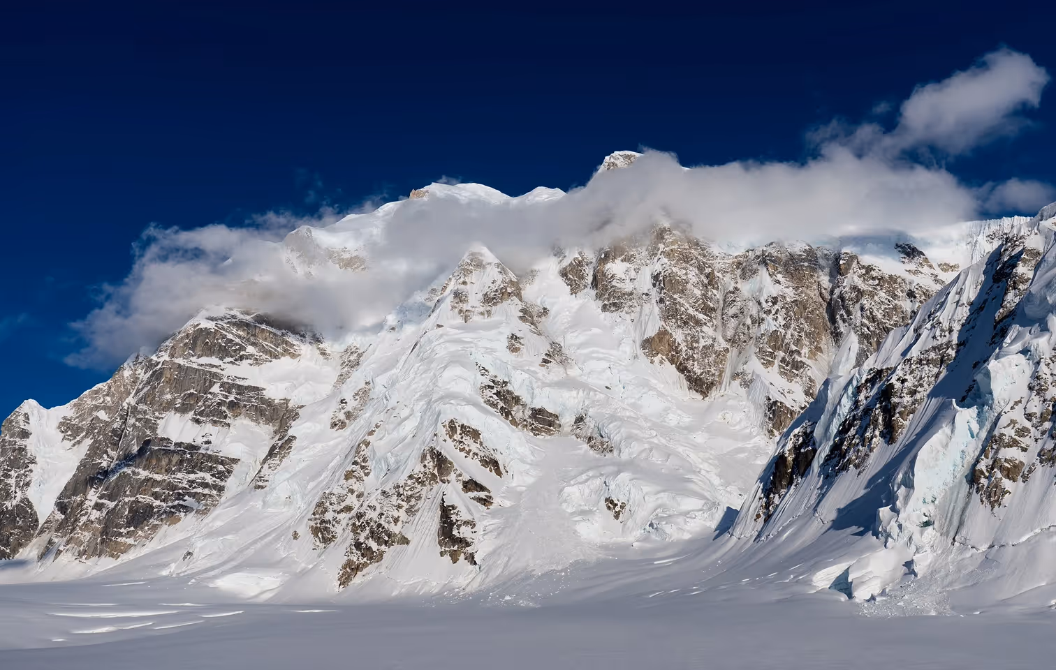 Snow-covered rugged mountain peaks partially obscured by low clouds against a deep blue sky.