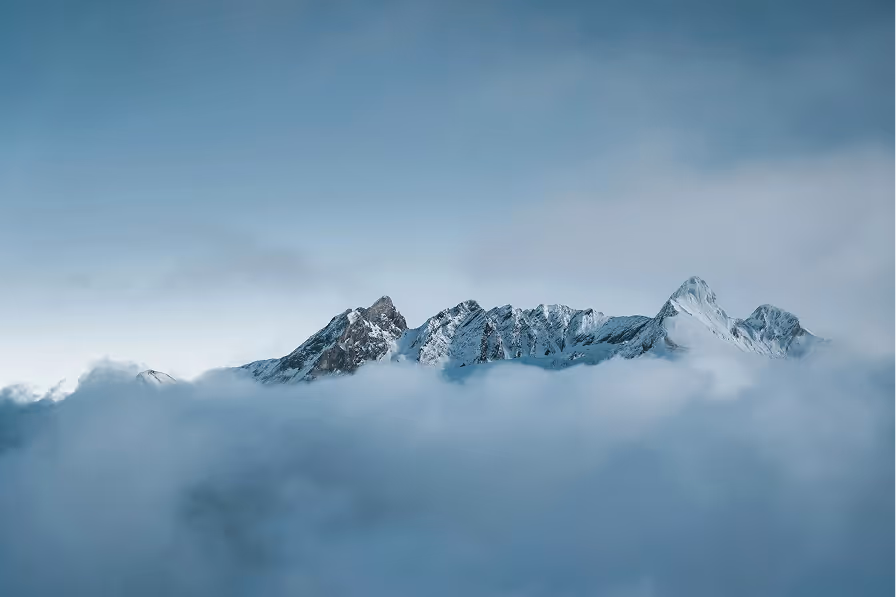 Snow-covered mountain peaks rising above thick clouds under a pale blue sky.