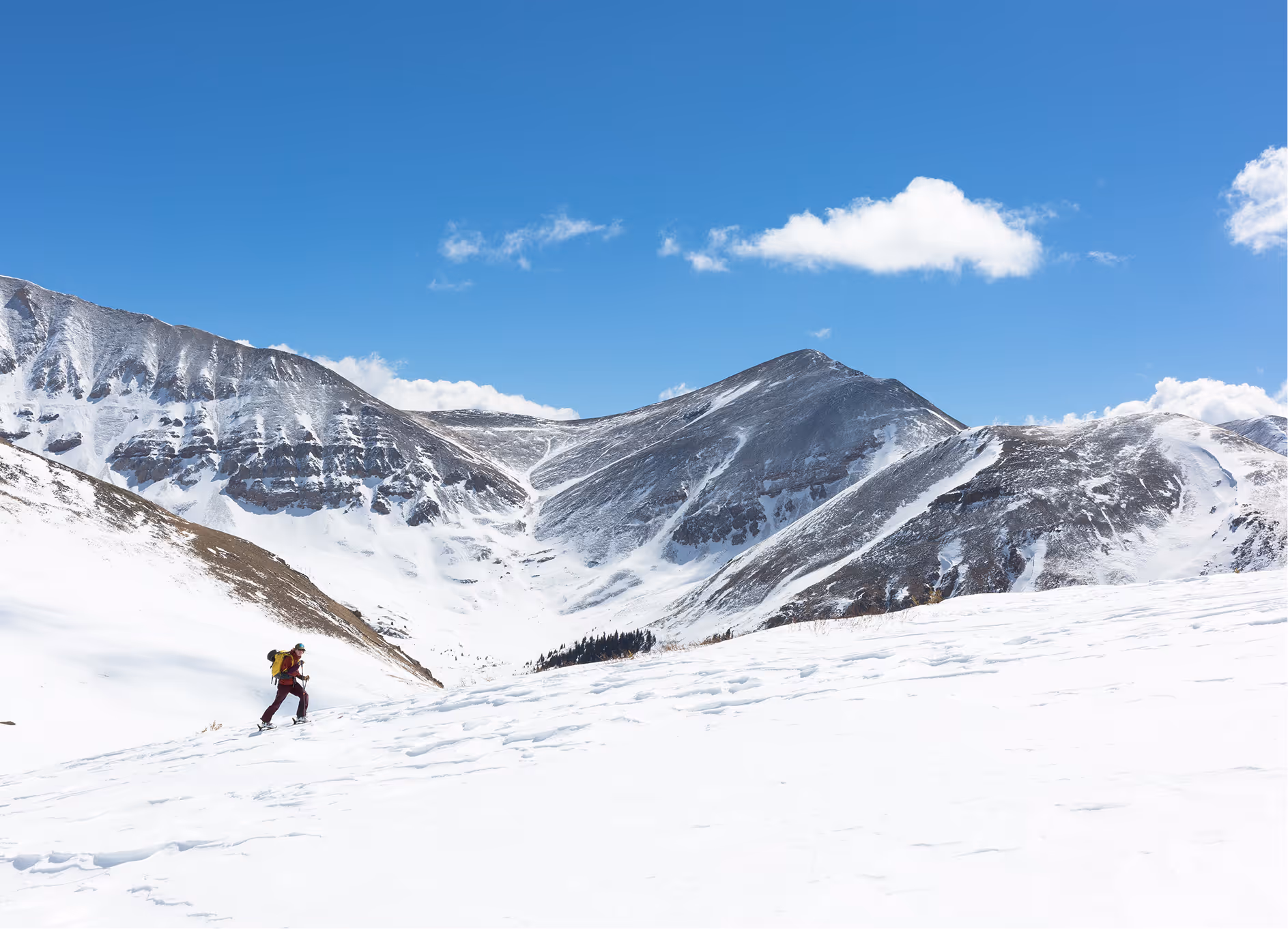 A person hiking uphill on a snowy mountain slope under a clear blue sky with scattered clouds.