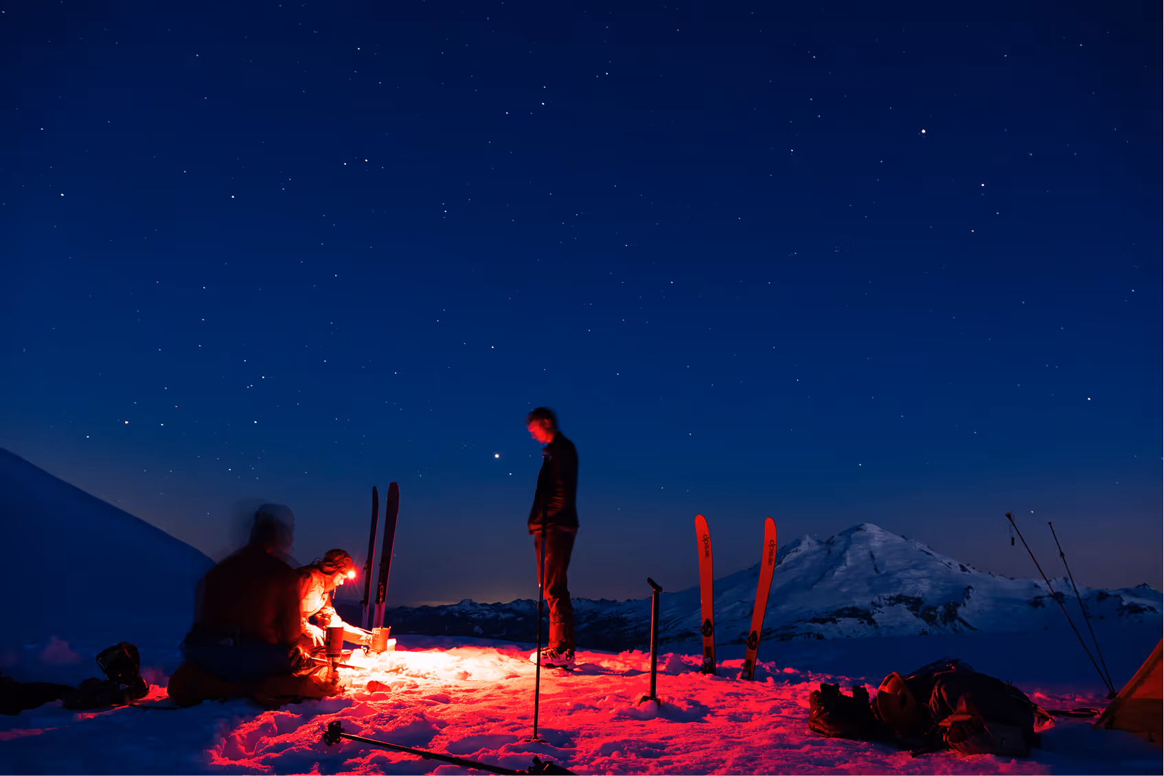 Three skiers on a snowy mountain illuminated by red light under a starry night sky with skis planted in the snow and a mountain in the background.