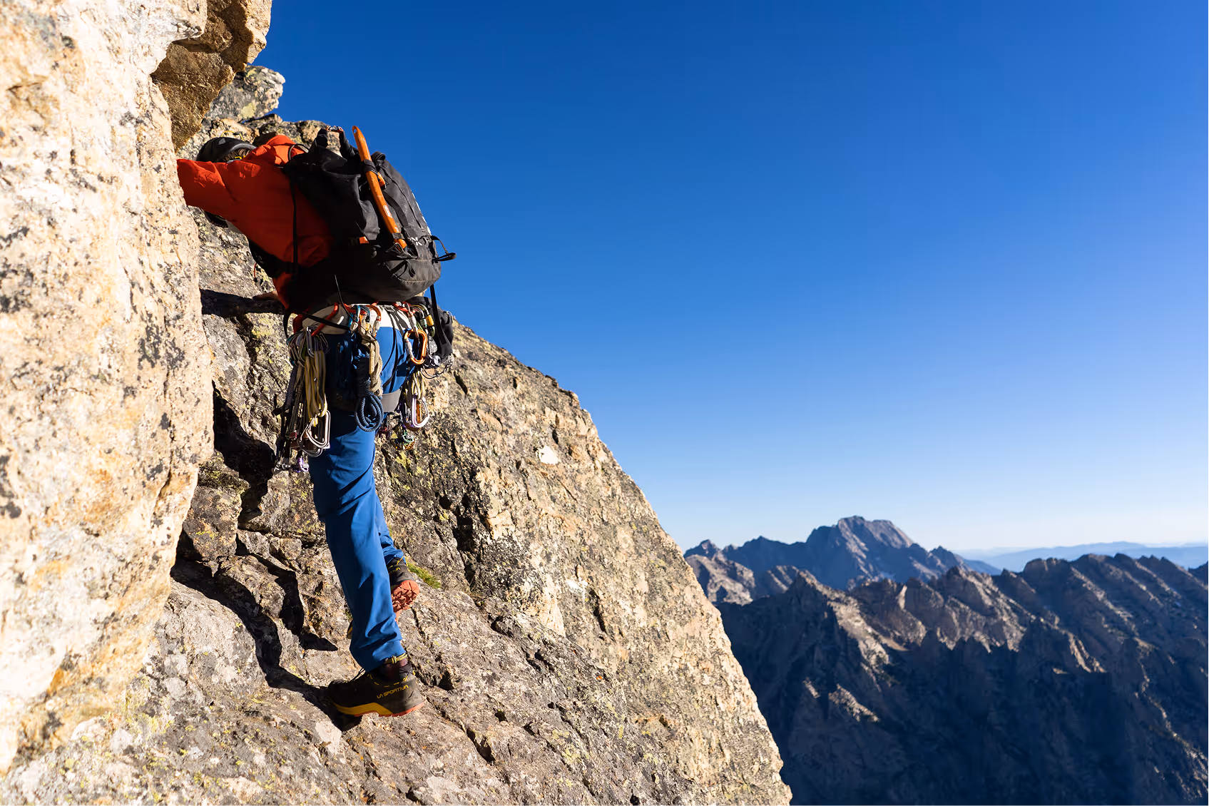 Climber in red jacket and blue pants ascending a rocky mountain face with climbing gear and a clear blue sky.