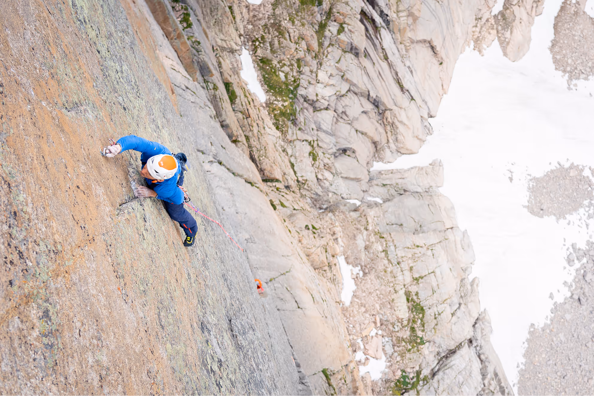 Climber wearing a white helmet and blue jacket ascending a steep rocky cliff with snow patches below.