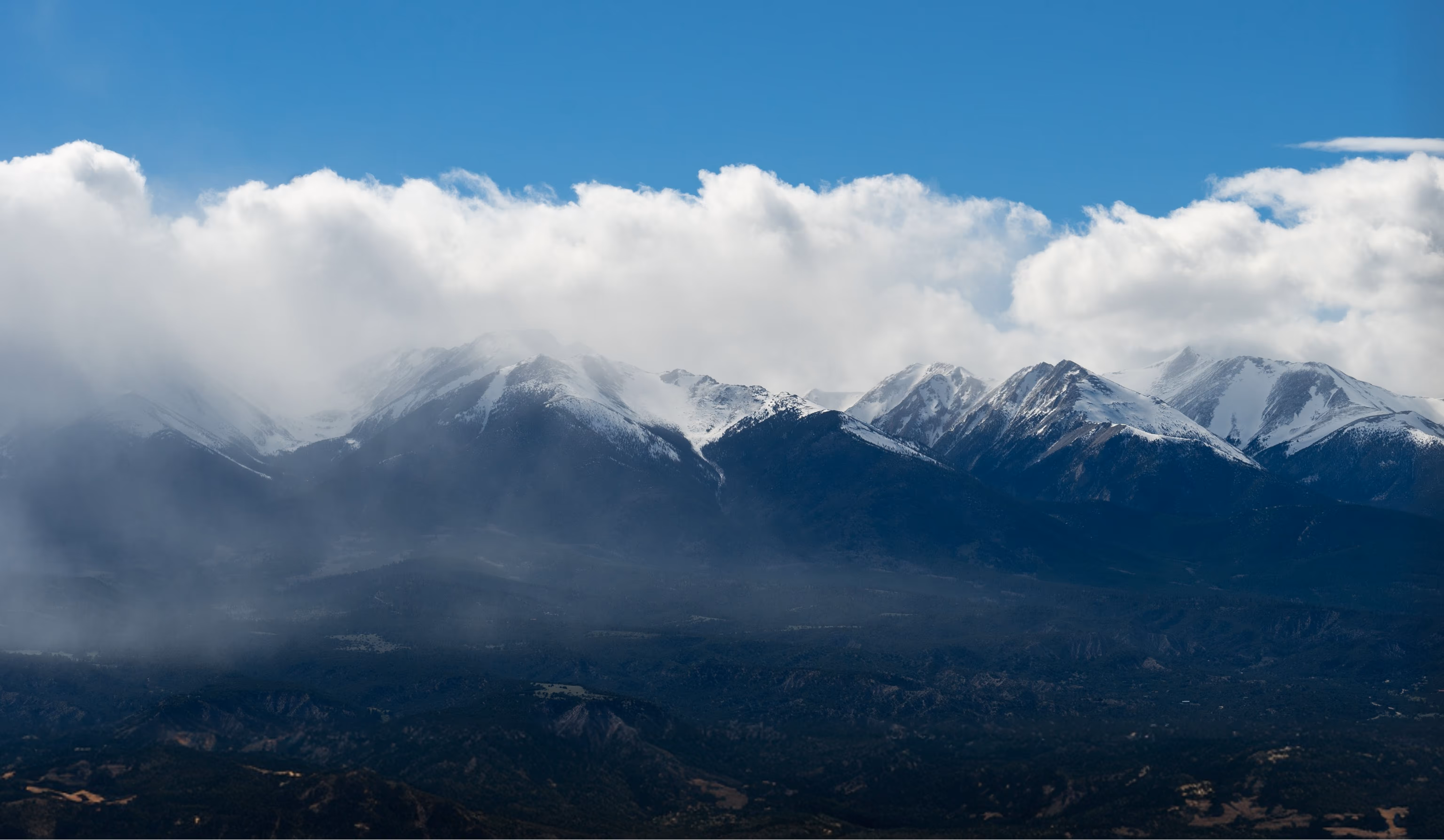 Snow-capped mountain range partially covered by dense fog under a blue sky with scattered clouds.